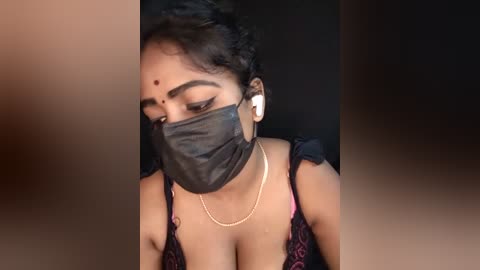 Media: Video of a South Asian woman with medium skin tone, short black hair, wearing a black face mask, pink and black patterned top, and white earbuds, set against a dark background.