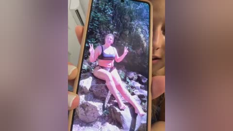 Media: Video of a woman in a black bikini sitting on a rocky beach, with a smartphone displaying a different photo of a woman in a bikini, held by a person's hand.