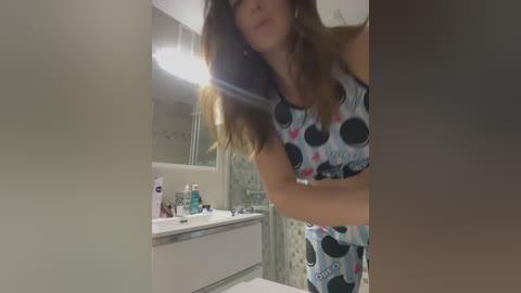 Video of a woman with long brown hair, wearing a blue and black polka-dot sports bra and matching leggings, in a bathroom with a white sink, toothbrush holder, and shower curtain.