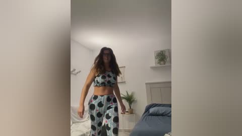 Media: Video of a woman with long brown hair wearing a matching polka dot sports bra and leggings, standing in a brightly lit bedroom with white walls, a bed, and potted plants.