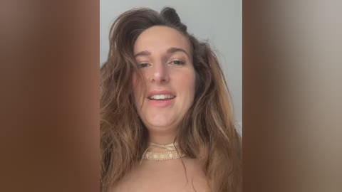 Media: Video of a smiling young woman with wavy brown hair and fair skin, wearing a cream-colored choker necklace. The background is blurred with soft, neutral tones.