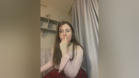 Media: Video of a young woman with long brown hair, fair skin, and blue eyes, wearing a light pink long-sleeve top and red plaid skirt, biting her finger, in a room with white walls, gray curtains, and a shelf with decorative items.