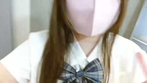 Media: Video of a person wearing a white shirt with a blue plaid bow tie, partially covered by a pink surgical mask, blurred background.