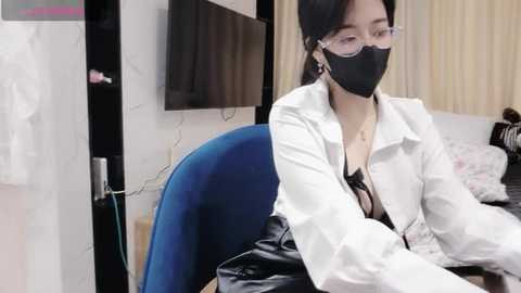 Media: Video of an East Asian woman with long black hair, wearing a white lab coat, black face mask, and glasses, seated at a desk in a clinical room with a blue chair and white walls.