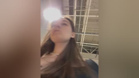 Media: Video of a young woman with long brown hair, wearing a white top, taking a selfie in a dimly lit room with metal bars in the background.