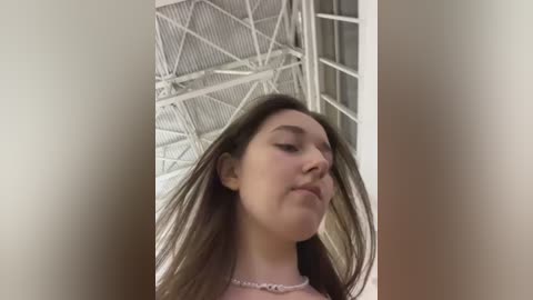 A video of a young woman with long, straight brown hair, fair skin, and a pink necklace, standing indoors with a white, metallic ceiling and window in the background.