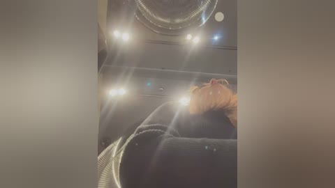 Video of a person with light brown hair wearing a dark sweater, lying on a circular fan, with bright lights illuminating the scene, creating a surreal, almost dreamlike atmosphere.