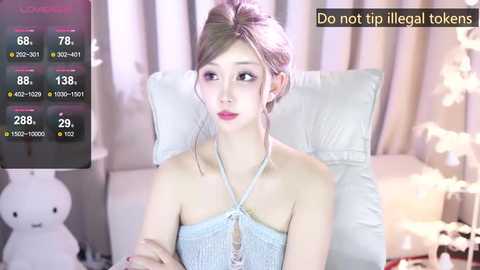 Media: A video of a young Asian woman in a light blue halter top, sitting on a white leather chair in a softly lit room with a teddy bear.