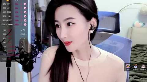 Media: Video of an East Asian woman with long black hair, red lipstick, wearing headphones and a white top, sitting in a hospital room with medical equipment.