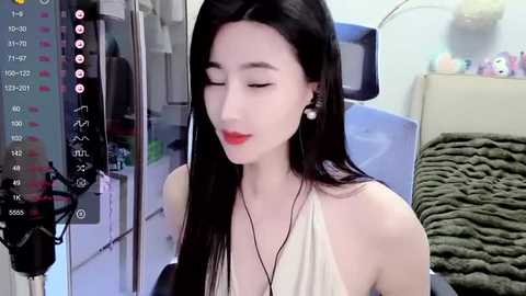 Video of a slender, East Asian woman with long black hair and fair skin, wearing a beige sleeveless top, sitting in a modern, well-lit room with a TV, green blanket, and medical equipment on the left.