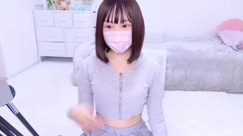 Media: Video of an East Asian woman with straight brown hair, wearing a light pink face mask and light grey zip-up top, sitting on a white floor in a modern bedroom with light grey furniture.