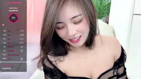 Media: A video of an Asian woman with light skin and shoulder-length dark hair, wearing a black lace bra, smiling. The background includes a green plant and a white surface.