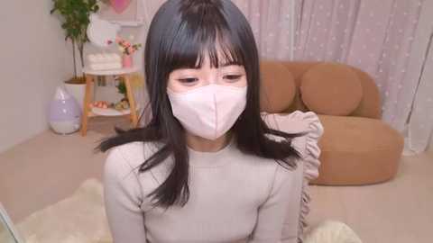 Media: Video of an East Asian woman with straight black hair, wearing a white mask, beige sweater, and beige mask, seated indoors with a beige sofa and a potted plant in the background.