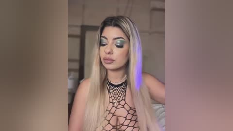 Video of a light-skinned, blonde-haired woman with long hair, wearing a black fishnet top revealing ample cleavage, and blue-green eye makeup, standing indoors with a blurred background.