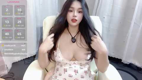 Media: A video of an Asian woman with long black hair and fair skin, wearing a revealing floral dress, sitting on a white chair. Background features white curtains and a virtual reality display.