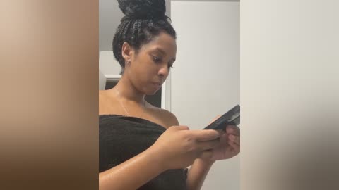 Video of a Black woman with light brown skin and short, tightly coiled hair wrapped in a black towel. She's focused on her smartphone, standing in a minimalist, modern bathroom with white walls.