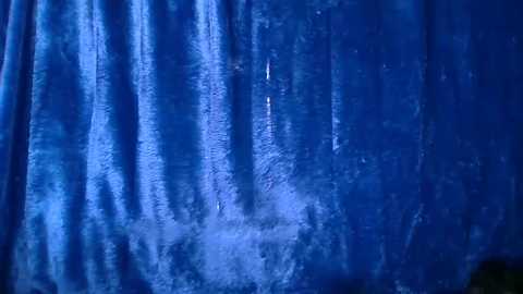 Media: Video of a textured, blue fabric backdrop with vertical pleats, slightly reflective, suggesting a stage or event setting. The fabric appears smooth but has subtle wrinkles, adding depth and dimension.