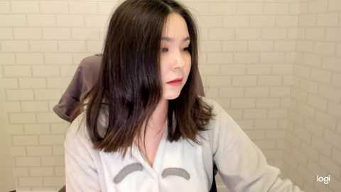 Video of a young East Asian woman with shoulder-length, straight, dark brown hair, wearing a white shirt with a gray elephant graphic. She stands in a white-tiled bathroom, looking down thoughtfully.