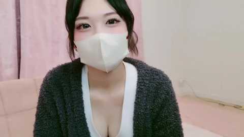 Media: Video of an East Asian woman with fair skin, wearing a dark knit cardigan over a low-cut white top, revealing cleavage, and a white face mask. She has dark hair pulled back and sits on a light-colored sofa in a softly lit room with pink curtains.