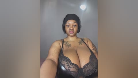 Video of a plus-sized Black woman with dark skin and short black hair, wearing a black lace bra that accentuates her large breasts. She has tattoos on her chest and arms. The background is a blurred, dimly lit room with a bright ceiling light.