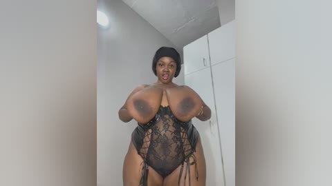 Video of a voluptuous, dark-skinned woman with a short black bob hairstyle, wearing a revealing black lace bodysuit, standing in a minimalist room with white walls and a door.