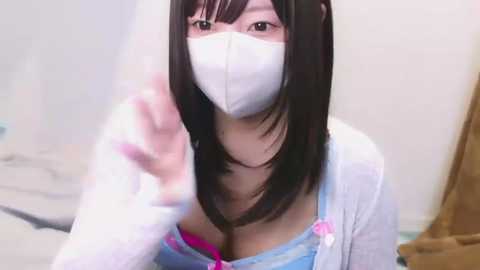 Video of an East Asian woman with long black hair, wearing a white face mask and pink bra under a light blue top. She gestures with her right hand, in a modern indoor setting.