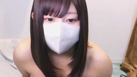 Video of an East Asian woman with long, straight brown hair, wearing a white surgical mask, sitting on a bed with a light-colored sheet. She has a slender build and light skin tone.