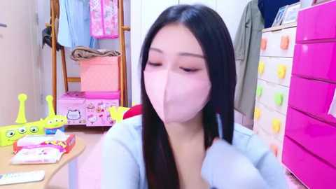 Media: Video of a young Asian woman with long black hair, wearing a pink face mask, seated in a colorful, cluttered room filled with toys, clothes, and storage bins.