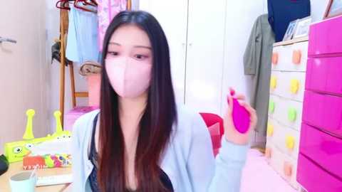 Media: Video of an East Asian woman with long, straight black hair and pale skin, wearing a pink mask, white cardigan, and black top, sitting in a colorful, organized room with pink drawers, toy shelves, and a wooden chair.