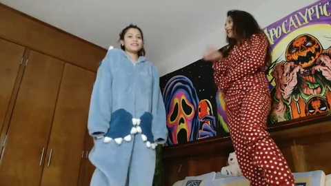 Video of two women in Halloween costumes: one in a blue robe with white pom-poms, the other in a red polka-dot dress, in a room with colorful wall art and wooden cabinets.