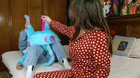 Media: Video of a woman in a red polka-dot pajama set, sitting on a bed, playing with a child in a blue, plush, Mickey Mouse costume. The room has wooden paneling and colorful, abstract wall art.