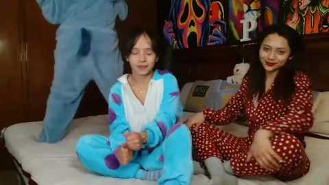 Video of two women in matching red polka-dot pajamas on a bed, one with a white fuzzy bear; another woman in blue pajamas stands behind them. Background features a colorful, abstract wall painting.