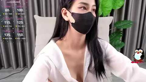 Media: Video of a young Asian woman with long black hair, wearing a black face mask, white shirt, and gray curtains in the background.