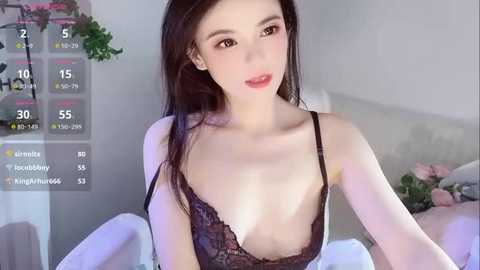 Video of a slender, fair-skinned Asian woman with long dark hair, wearing a black lace bra, sitting on a bed with white sheets, in a softly lit room with greenery and flowers in the background.