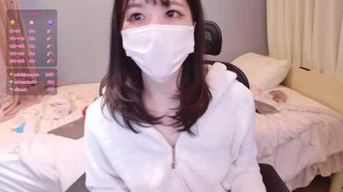 Media: A video of an Asian woman with long, dark hair, wearing a white surgical mask and robe, sitting on a bed in a cozy room.