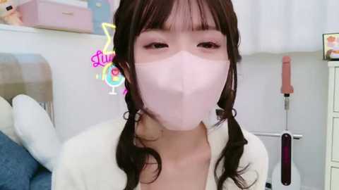 Media: Video of an Asian woman with long dark hair in pigtails, wearing a white mask, pink lipstick, and a white jacket, in a cozy room with pastel decor and plush toys.