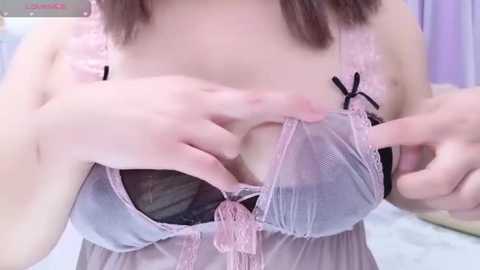 Media: A video of a woman with light skin, wearing a sheer, pink and black lingerie bra, adjusting it with her hands. Background includes a bed and blurred purple curtains.