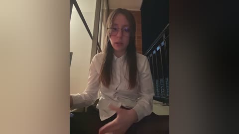 Video of a young woman with glasses, long brown hair, wearing a white button-up shirt, sitting on a bed, holding a phone, dimly lit room.