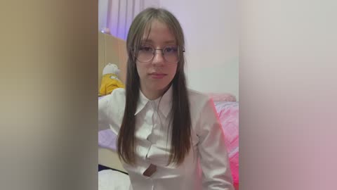 Media: Video of a young Caucasian woman with straight, long brown hair, wearing glasses and a white shirt, sitting in a dimly lit room with pinkish-purple walls, a bed with white sheets, and a yellow jacket hanging on a chair.