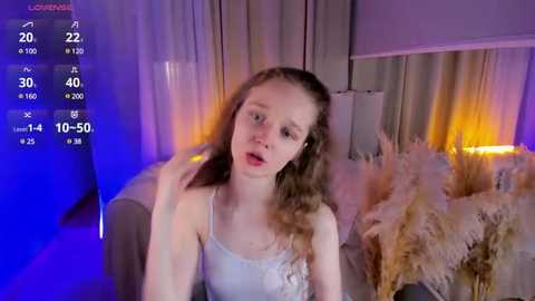 Media: A video of a fair-skinned, curly-haired woman in a white top, sitting on a couch with beige cushions and dried flowers, in a dimly lit room with purple and blue lighting.