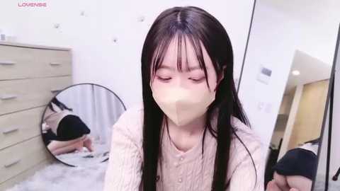 Video of a young woman with long black hair, wearing a beige face mask, sitting on a white carpeted floor in a modern bedroom with a wooden dresser and mirror.