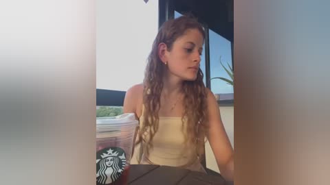 Media: Video of a young Caucasian woman with long, wavy brown hair, wearing a beige tank top and sitting in a Starbucks, holding a large cup.