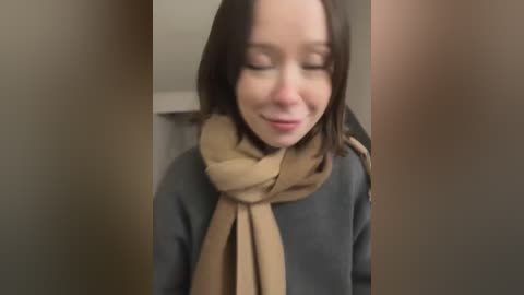 Media: Video of a young woman with shoulder-length brown hair, closed eyes, and a beige scarf tied around her neck, smiling gently. She wears a dark gray sweater. Background is blurred, suggesting indoor setting.