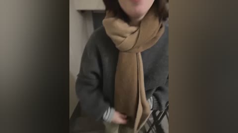 Media: Video of a person with shoulder-length brown hair, wearing a grey sweater and a tan scarf, standing in a dimly lit hallway with beige walls. The image is slightly out of focus.