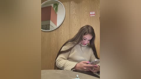 Media: Video of a young woman with long brown hair, wearing a cream sweater, reading a phone in a beige wooden room with a circular mirror and plant.