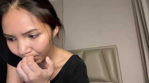 Media: A video of an East Asian woman with long black hair, wearing a black top, biting her finger, standing in a minimalist room with beige walls and a tufted headboard.