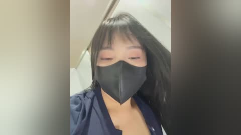 A video of an Asian woman with long black hair and fair skin, wearing a black face mask, navy shirt, and a visible bra strap, standing indoors with blurred background.