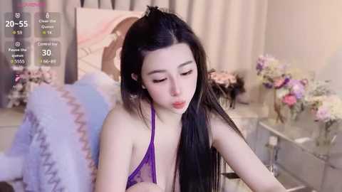 Media: Video of a fair-skinned, slender Asian woman with long black hair, wearing a purple lace bra, sitting in a cozy room with a bed, flowers, and a TV.