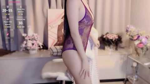 Media: A video shows a fair-skinned woman in a sheer, purple lace bodysuit, standing in a softly lit bedroom with pale pink flowers, a white dresser, and a pink painting.