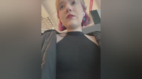 Video of a young woman with pale skin and pink hair, wearing a black halter top, sitting in a car. Background shows blurry interior with a brick wall.